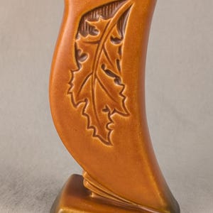 May include: A tall, curved ceramic vase with a glossy, warm amber-brown glaze. The front surface features a deeply embossed, detailed oak leaf design. The base is wide and stable, transitioning into a slender, upright body with a narrow opening.