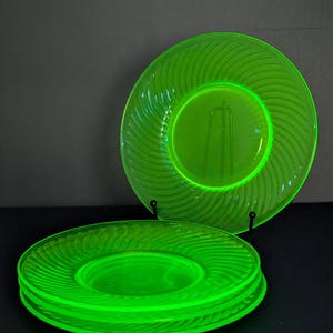 Può includere: Un set di quattro piatti in vetro verde neon vibrante caratterizzati da un distinto motivo radiale a spirale sul bordo. I piatti sono traslucidi e circolari, con uno esposto in verticale su un supporto nero e tre impilati in piano.