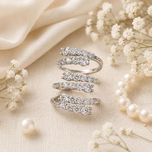 May include: A silver-toned bypass ring featuring a spiral design adorned with multiple round, clear, faceted stones. The ring is positioned among small white flowers and several smooth, round, cream-colored pearls on a textured fabric surface.