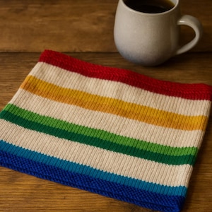 May include: A knitted neck warmer with horizontal stripes in red, yellow, green, and blue. The neck warmer is made of a soft, textured material. A gray mug filled with coffee sits in the background on a wooden surface.