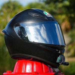 Op de afbeelding: Een matzwarte integraalhelm voor motorrijders met een reflecterend zilverkleurig vizier en een AGV-logo aan de voorzijde. De helm is voorzien van een zwarte kinband en een kleine zwarte camerabevestiging aan de rechterkant van de kin.