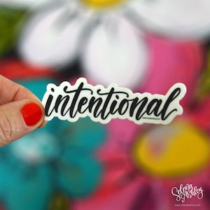 May include: A white sticker with the word "intentional" written in black cursive script. The sticker is held in a hand against a colorful background.