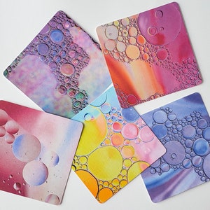 May include: Five square coasters with abstract designs featuring colorful, iridescent bubbles. The coasters are made of a thick, durable material and are perfect for protecting surfaces from spills and scratches.