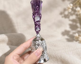 May include: A decorative hand bell featuring a silver-toned metal base with intricate embossed patterns. The handle is topped with a faceted purple amethyst crystal, accented by a swirling purple wire wrap and a small, clear spherical stone.