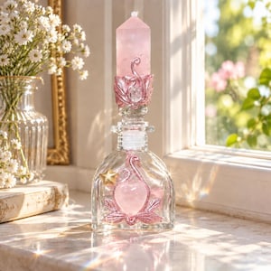 May include: A clear glass bottle with a stopper topped by a pink rose quartz crystal point. The bottle features a pink metallic leaf-shaped base, a central pink heart-shaped stone, a small gold star, and decorative swirling metallic accents.