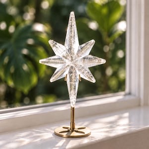 May include: A clear, multi-pointed star ornament with a textured, crystalline interior. The star is mounted on a thin, vertical brass-colored metal rod attached to a circular, flat base with four symmetrical cutouts in a floral pattern.