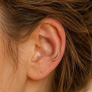 May include: A curved gold ear climber earring featuring a row of small, round, clear stones set along the arc. The piece is positioned along the inner cartilage of the ear, following the natural curve of the ear shape with a delicate design.