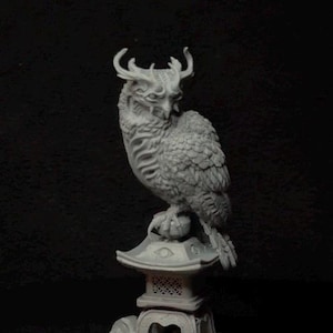 May include: A light gray, unpainted resin figurine of a horned owl perched atop a traditional stone lantern. The owl features detailed feathers and curved horns, while the lantern base rests on a circular platform with a textured ground surface.