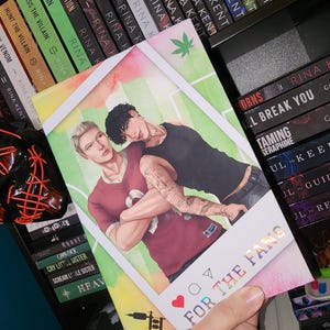 May include: A rectangular card featuring a digital illustration of two individuals embracing, one with blonde hair in a maroon shirt and one with dark hair in a black tank top. The card includes a white border, social media icons, and the text FOR THE FANS.