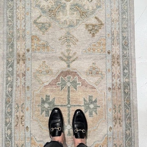 May include: A rectangular area rug featuring a faded, intricate geometric pattern in muted shades of sage green, dusty rose, and golden tan on a cream-colored background, with a decorative border and a textured, woven pile construction.