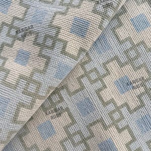 May include: A woven rug featuring a repeating geometric pattern in light blue, sage green, and cream. The design consists of interconnected green lines forming square shapes with blue centers. The text KARIBAR RUGS is printed on the surface.