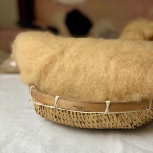 May include: A round, shallow woven bamboo basket filled with a large, fluffy mound of raw, light tan wool fiber. The basket features a thin, smooth wooden rim secured with small, light-colored ties, holding the soft, textured wool in place.