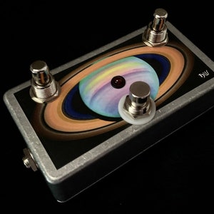 May include: A silver effect pedal with a colourful image of a planet with rings. The pedal has three buttons, one red LED, and the text "bw" on the side.