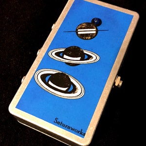 May include: A silver effect pedal with a blue background and three black and white planets with rings. The text "Saturnworks" is printed on the bottom of the pedal.