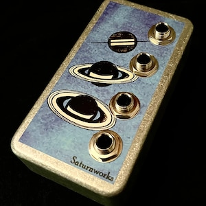Puede incluir: Un pedal de efectos de guitarra dorado y azul con cuatro conectores dorados y el texto "Saturnworks" en la parte inferior. El pedal tiene un diseño de temática espacial con planetas y anillos.