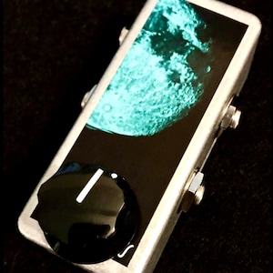 Puede incluir: Un pedal de efectos de guitarra rectangular plateado con un gráfico de una luna color verde azulado sobre un fondo negro. El pedal incluye una perilla de control negra grande con una línea indicadora blanca y dos conectores de metal.