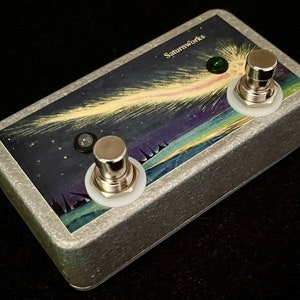 May include: A silver-colored Saturnworks effects pedal with a cosmic design. The pedal features a painted illustration of a comet streaking across a starry night sky. It has two silver footswitches and two indicator lights. The text "Saturnworks" is printed on the top.