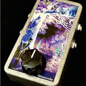 以下が含まれることがあります： 森の中にいる女の子と猫のカラフルなグラフィックが描かれた銀色のエフェクトペダル。ペダルには黒いノブとトグルスイッチが付いています。ペダルの底には「Saturaworks」という文字が印刷されています。