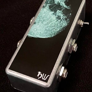 May include: A silver metal effects pedal with a black and white image of the moon on the top. The pedal has two 1/4 inch jacks and a toggle switch on the side. The pedal is labelled "bw".