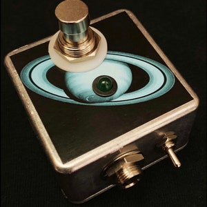 以下が含まれることがあります： 上面が黒と青の土星の絵柄の銀色のフットペダル。ペダルには銀色のボタンと緑色のLEDライトが付いている。