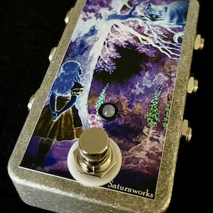 Puede incluir: Una caja de pedales plateada con una imagen colorida de una niña y un gato en un bosque. La caja tiene cinco botones y un botón grande con un anillo blanco. El texto "Saturnworks" está impreso en la parte inferior de la caja.