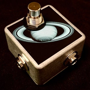 May include: A silver foot pedal with a black and white image of Saturn on the top. The pedal has a silver button and two silver jacks on the sides.
