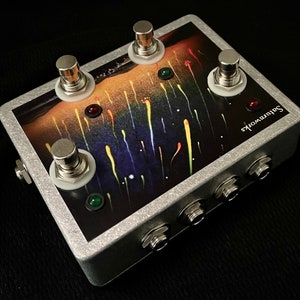 May include: A silver effects pedal with three footswitches and four jacks. The pedal has a colourful graphic design with a black background and a yellow, orange, and red gradient.