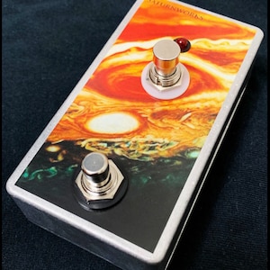 May include: A silver stompbox with a graphic of Jupiter and its red spot. The box has two footswitches, one with a red LED and one with a black button.