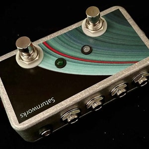 May include: A silver stompbox with a black and blue graphic design featuring a red line and a green LED light. The stompbox has two footswitches and four jacks. The text "Saturnworks" is printed on the side.