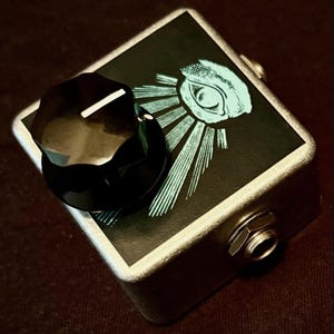 Peut inclure: Pédale d'effets argentée avec un dessus noir orné d'un motif d'œil bleu clair. La pédale est dotée d'un gros bouton noir et d'une prise d'entrée argentée. Le motif est un œil stylisé avec des lignes rayonnantes.