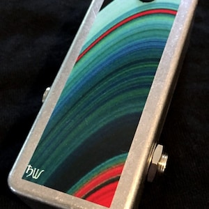 May include: A silver metal effects pedal with an abstract design in blue and green, featuring a red stripe. The pedal has a single footswitch and a blue LED. The pedal is labelled "hw".