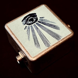 Puede incluir: Una pequeña caja cuadrada de color dorado con una imagen en blanco y negro de un ojo con rayos de luz que emanan de él. La caja tiene dos conectores metálicos en el lateral.