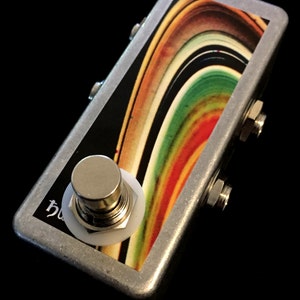 以下が含まれることがあります： 上面がカラフルな抽象的なデザインの銀色のストンプボックス。ストンプボックスには、白いボタンが付いたフットスイッチが1つと、側面に1/4インチのジャックが2つあります。