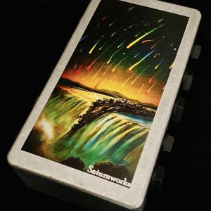 Może przedstawiać: Srebrne metalowe pudełko z kolorowym obrazkiem wodospadu i nocnego nieba z gwiazdami spadającymi. Pudełko ma napis "Saturnworks" nadrukowany na spodzie.