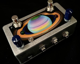 Puede incluir: Un pedal de efectos plateado con una imagen colorida de un planeta con anillos. El pedal tiene dos interruptores de pie, una perilla y tres conectores.