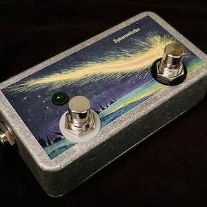 May include: A silver stompbox with two footswitches and a green LED. The stompbox features a graphic of a comet in a starry night sky. The text "Saturnworks" is printed on the top of the stompbox.
