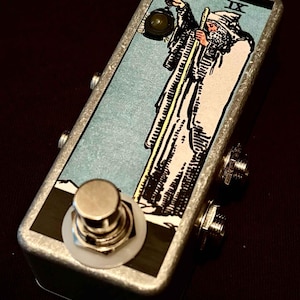 Könnte beinhalten: Ein silbernes Effektpedal aus Metall mit einem Schwarz-Weiß-Tarotkarten-Design mit der IX der Stäbe. Das Pedal hat einen einzigen Fußschalter und zwei Buchsen.