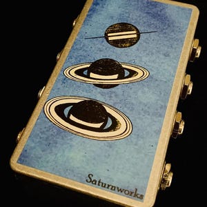 Może przedstawiać: Złote metalowe pudełko z niebieskim tłem i trzema czarno-białymi ilustracjami Saturna. Pudełko ma pięć złotych gałek po bokach i napis "Saturnworks" na dole.
