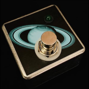 May include: A silver stompbox with a black and blue graphic of Saturn. The stompbox has a white button with a gold knob and a green LED light.