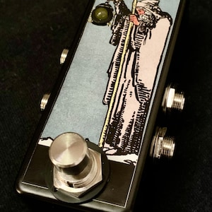 Pode incluir: Um pedal de efeitos de guitarra preto com um botão prateado e duas tomadas de entrada. O pedal apresenta um design de carta de tarô com a imagem da carta do Eremita, que representa uma figura segurando uma lanterna e um cajado. O número da carta, "IX", é visível no canto superior direito.