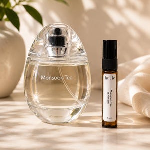 May include: A clear, oval-shaped glass perfume bottle labeled Monsoon Tea d'Annam next to a slender, dark amber glass vial with a black cap, labeled huele Monsoon Tea d'Annam 5 mL. Both containers feature white text on their respective surfaces.