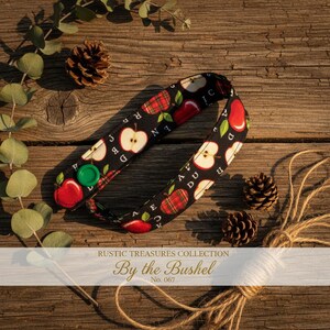 May include: A black fabric strap featuring a repeating pattern of red apples and green leaves, secured with a single bright green plastic snap button. The strap is printed with the text RUSTIC TREASURES COLLECTION By the Bushel No. 067.