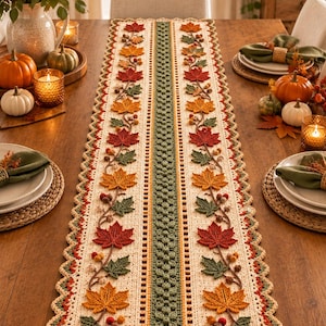 May include: A rectangular crochet table runner featuring a cream-colored base with a central green textured stripe. The runner is decorated with repeating patterns of crocheted autumn maple leaves in red, orange, and green, accented with small brown berries.