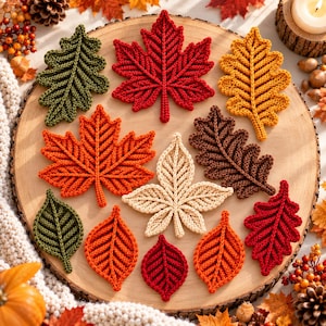 May include: A collection of twelve crocheted leaves in various shapes and autumn colors, including red, orange, yellow, brown, cream, and green. The leaves feature intricate stitched vein details and are arranged on a round wooden surface.
