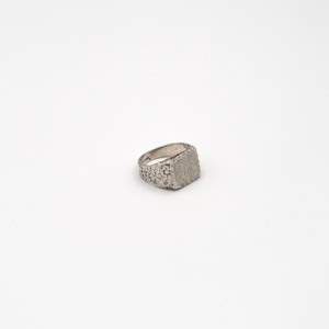 May include: A silver-toned signet ring featuring a textured, rugged surface across the band and the square-shaped face. The metal has a matte, granular finish that creates a consistent, weathered appearance throughout the entire piece of jewelry.
