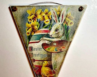 Può includere: Un cartello di legno triangolare con un'immagine vintage di Pasqua. Un coniglio bianco dipinge un uovo con un pennello e una tavolozza. Sono presenti anche narcisi gialli e un pulcino giallo. Il cartello recita "Easter Greetings".