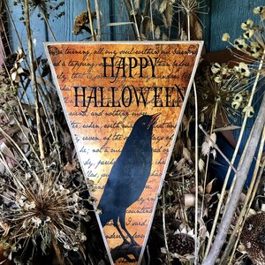 Puede incluir: Cartel triangular de Halloween con una silueta de cuervo negro y las palabras "HAPPY HALLOWEEN". El cartel tiene un aspecto vintage con un fondo de escritura y está rodeado de flores y follaje secos.