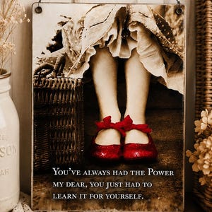 May include: A rectangular metal wall sign featuring a sepia-toned photograph of legs wearing bright red shoes with bows, next to a wicker basket. The text reads: "YOU'VE ALWAYS HAD THE POWER MY DEAR, YOU JUST HAD TO LEARN IT FOR YOURSELF."