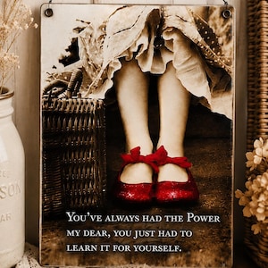 May include: A rectangular metal wall sign featuring a sepia-toned photograph of legs wearing bright red shoes with bows, next to a wicker basket. The text reads: "YOU'VE ALWAYS HAD THE POWER MY DEAR, YOU JUST HAD TO LEARN IT FOR YOURSELF."