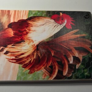 May include: Rectangular wooden cutting board featuring a detailed image of a rooster. The rooster has red, brown, and white feathers, with a red comb and wattle. A leather loop is attached for hanging. The background includes a chicken wire fence.